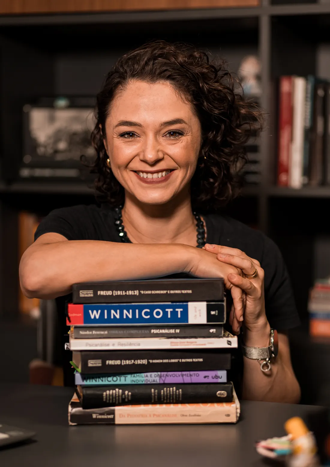 Bibiana Malgarim sorrindo, apoiada sobre uma pilha de livros de Freud e Winnicott